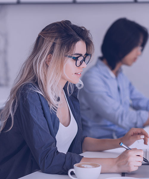 A young woman with blonde hair and glasses is focused on taking notes at a desk, while a man sits in the background working on a laptop.